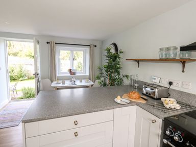 A kitchen with a countertop holding a loaf of bread toaster and eggs next to a dining table by a window and an open door at Dairy Cottage in Abbotsbury
