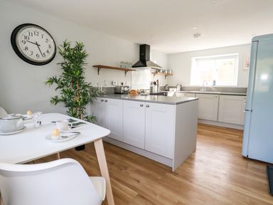 A kitchen with white cabinets wooden floor a dining table with chairs a large wall clock and a potted plant at Dairy Cottage in Abbotsbury