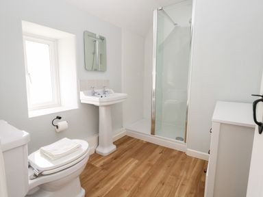 A bathroom with a toilet, pedestal sink, shower enclosure, window, and white cabinet at Dairy Cottage in Abbotsbury