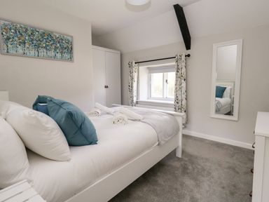 A bedroom with a white bed with blue pillows a window with curtains a white wardrobe and a wall mirror at Dairy Cottage in Abbotsbury