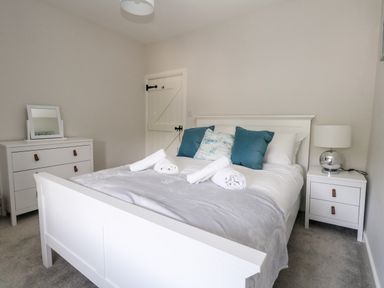 A bedroom with a white bed with blue pillows and rolled towels a dresser with a mirror and a nightstand with a lamp at Dairy Cottage in Abbotsbury
