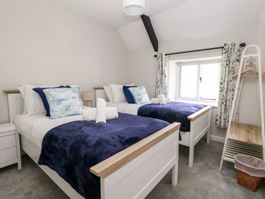 A bedroom with two single beds blue blankets pillows a window with floral curtains and a wooden shelf at Dairy Cottage in Abbotsbury