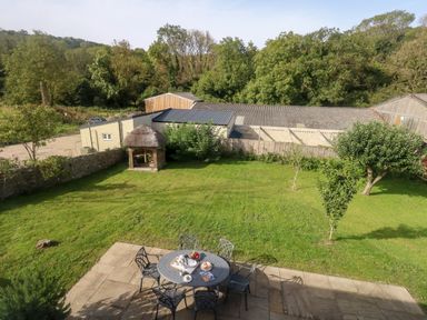 A garden with a round table and chairs on a patio and a thatched gazebo in the background at Dairy Cottage in Abbotsbury