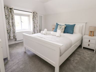 A bedroom with a white bed and blue pillows next to a window with floral curtains at Dairy Cottage in Abbotsbury