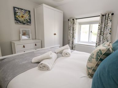 A bedroom with a bed with rolled towels pillows a wardrobe a chest of drawers with a mirror a window with floral curtains and a floral painting on the wall at Dairy Cottage in Abbotsbury