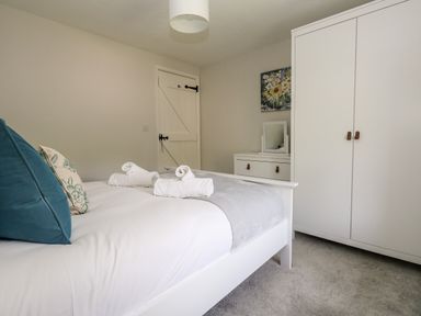 A bedroom with a white bed and folded towels a white wardrobe and a chest of drawers with a mirror and floral painting at Dairy Cottage in Abbotsbury