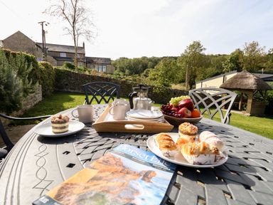 An outdoor metal table with cups plates pastries fruit and a magazine in a garden at Dairy Cottage in Abbotsbury