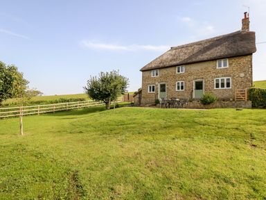 A stone cottage with a thatched roof and patio furniture on a lawn with trees at Dairy Cottage in Abbotsbury