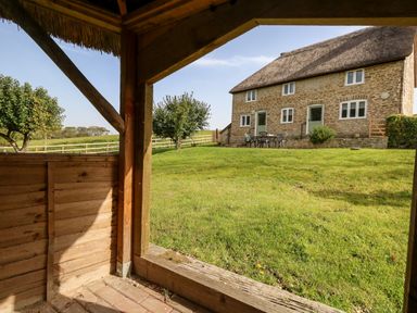 A stone house with a thatched roof and outdoor seating in a grassy yard with trees at Dairy Cottage in Abbotsbury