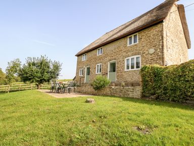 A stone house with a thatched roof and green doors with an outdoor patio set in a garden at Dairy Cottage in Abbotsbury
