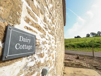 A stone wall with a Dairy Cottage sign and a rural landscape with a road and green fields at Dairy Cottage in Abbotsbury