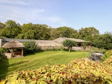 A garden with grass trees a thatched gazebo a table with chairs and farm buildings in the background at Dairy Cottage in Abbotsbury