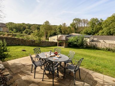 An outdoor patio with a metal table and six chairs with tea and snacks on the table in a garden with trees and a wooden gazebo at Dairy Cottage in Abbotsbury