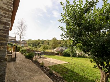An outdoor patio area with a table and chairs on a stone patio next to a grassy garden with trees and a thatched gazebo at Dairy Cottage in Abbotsbury