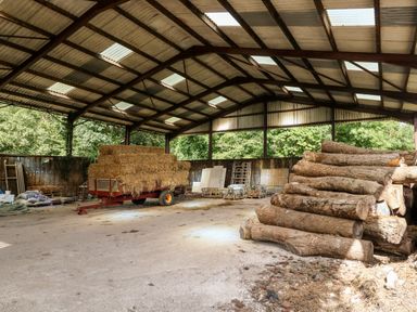 An open barn with stacked hay on a trailer and a pile of large logs inside at Dairy Cottage in Abbotsbury