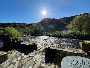 A stone patio beside a flowing river with trees and hills under a bright sun at Riverside Cottage in Beddgelert