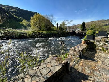 A stone patio by a flowing river with trees and hills in the background at Riverside Cottage in Beddgelert