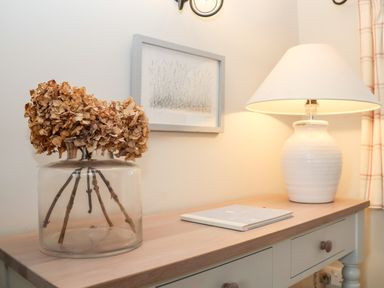 A wooden table with a glass vase containing dried flowers a book and a white table lamp on it at 2 Mosses Cottage in Friston