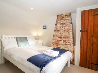 A bedroom with a white bed a blue pillow and blanket a brick chimney breast and wooden door at 2 Mosses Cottage in Friston