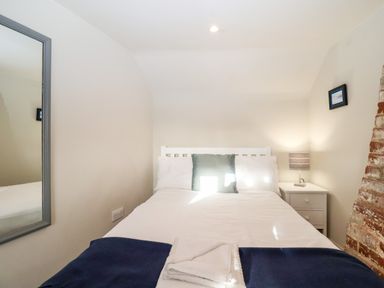 A bedroom with a bed white bedding a green pillow a bedside table with a lamp a mirror on the wall and an exposed brick section at 2 Mosses Cottage in Friston