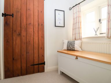 A wooden door next to a white bench with a cushion under a window with curtains at 2 Mosses Cottage in Friston