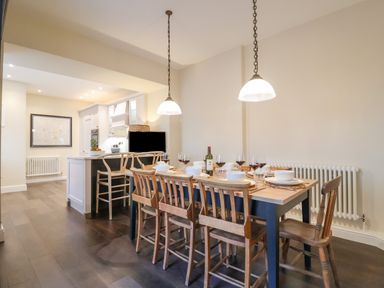 A dining area with wooden chairs around a table set with bowls and glasses next to a kitchen island with bar stools at The Middle House in Southwold