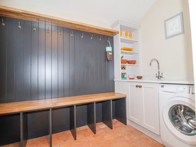 A utility room with wooden bench, coat hooks, shelves with bowls and glasses, sink and washing machine at The Middle House in Southwold
