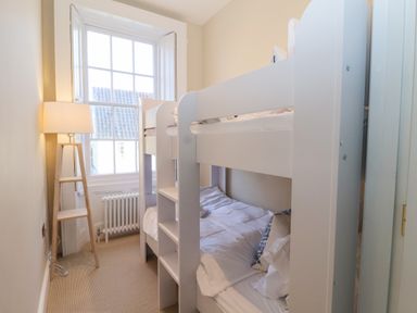 A small bedroom with a white bunk bed a wooden floor lamp and a window showing outside roof tiles at The Middle House in Southwold