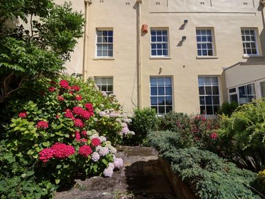 A garden with blooming flowers and bushes in front of a beige building with white-framed windows at The Middle House in Southwold