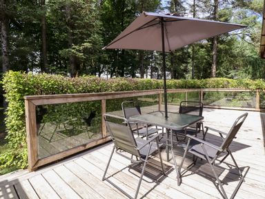 An outdoor wooden deck with a glass table and four chairs under a large umbrella surrounded by trees and hedges at Washburn Lodge Fewston near Darley