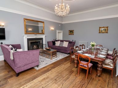 A living room with purple sofas a wooden dining table set with plates and glasses a fireplace and a chandelier at The Stables and West Wing Bolton Percy near Tadcaster