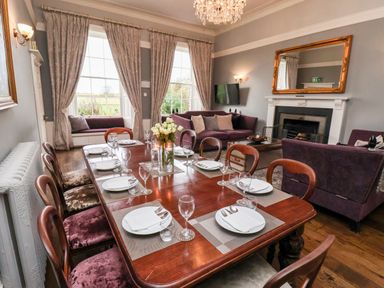 A dining room with a table set for eight with plates glasses and cutlery and purple sofas near a fireplace with a mirror above at The Stables and West Wing Bolton Percy near Tadcaster