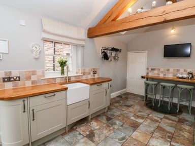 A kitchen with tiled floor wooden countertops a white farmhouse sink stools under a breakfast bar and a wall-mounted television at The Stables and West Wing Bolton Percy near Tadcaster