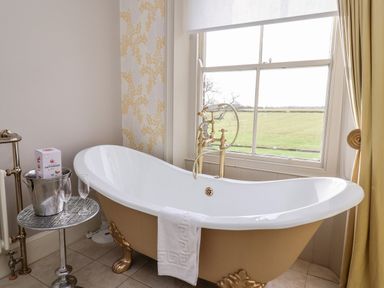 A bathroom with a clawfoot bathtub next to a window and a small table holding a champagne bottle and glasses at The Stables and West Wing Bolton Percy near Tadcaster