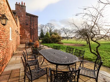 An outdoor patio with metal tables and chairs next to a brick building and a grassy area with trees at The Stables and West Wing in Bolton Percy near Tadcaster