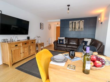 A living room with a brown leather sofa yellow chair wooden sideboard and a dining table with a bottle of wine and fruit at Pointers View in Weymouth