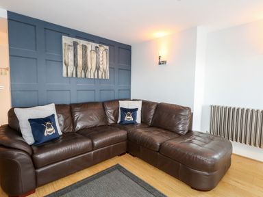 A living room with a brown leather sectional sofa and blue cushions in front of a blue paneled wall with wall art at Pointers View in Weymouth