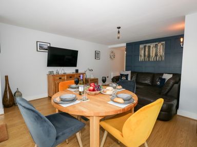A dining area with a round wooden table set for four and colorful chairs next to a living room with a dark sofa and TV at Pointers View in Weymouth