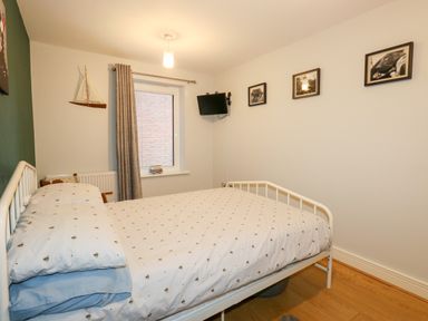 A bedroom with a bed covered in white bedding with patterns a small wall-mounted television a window with grey curtains three framed pictures on the wall a model sailboat on a shelf and wooden flooring at Pointers View in Weymouth