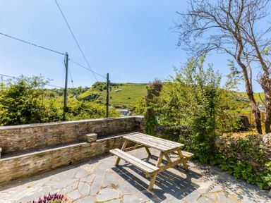 A stone patio with a wooden picnic table surrounded by plants and a stone wall with a view of green hills at Mermaid Cottage in Port Isaac