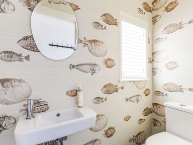 A bathroom with fish wallpaper a white sink with a faucet a soap dispenser a circular mirror a window with blinds and a white toilet at Mermaid Cottage in Port Isaac