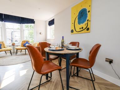A dining area with a round wooden table and four brown chairs beside a living room with yellow chairs and large windows at Flat 6 Thornlow Heights in Weymouth