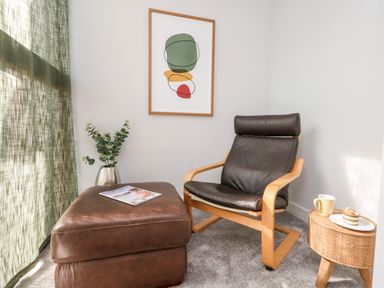 A corner with a leather chair next to a brown ottoman a small round side table with a cup and pastry and a framed abstract print on the wall at Flat 6 Thornlow Heights in Weymouth
