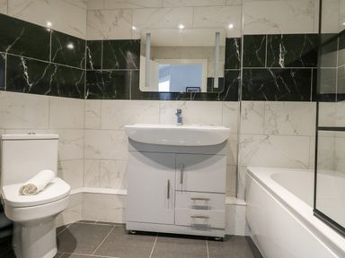 A bathroom with a toilet with a rolled towel on the lid a sink with a cabinet a mirror above the sink and a bathtub with a glass screen at Flat 6 Thornlow Heights in Weymouth