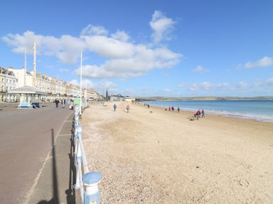 A sandy beach with people walking near the shoreline and buildings along the promenade at Flat 6 Thornlow Heights in Weymouth
