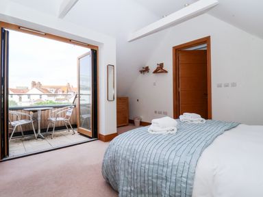 A bedroom with a bed with folded towels and a light blue blanket open doors leading to a balcony with two chairs and a table at The Old Gymnasium in Southwold