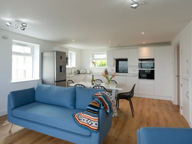 An open plan kitchen and living area with blue sofas a dining table with chairs and white kitchen cabinets at Treen in St Ives