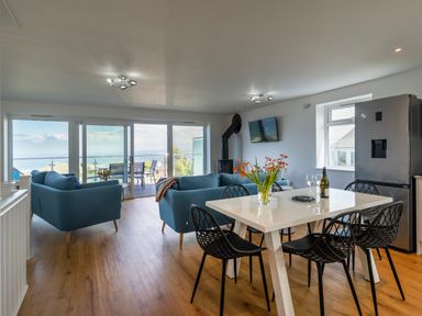 An open living and dining area with blue sofas a white dining table with black chairs a refrigerator and a wall-mounted TV with sea view at Treen in St Ives