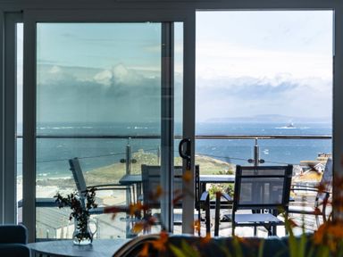 A balcony with chairs and a table overlooking the sea through glass doors at Treen in St Ives