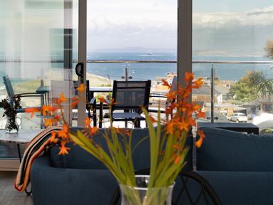 A room with a blue couch orange flowers and a view of a balcony with chairs and the sea beyond at Treen in St Ives